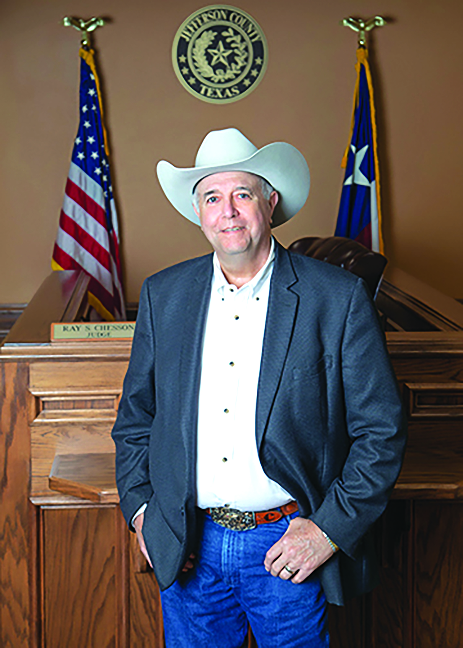 A photo of Ray Chesson. He is wearing jeans, a brown belt, a white button down shirt, and a grey jacket. He is wearing a white cowboy hat. 