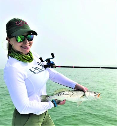 Taylor Garcia poses with a trout. She has a green neck scarf and green pants and a white long sleeve athletic shirt. She has a green hat with red text and sunglasses on. There is a fishing pole under har left arm.