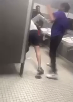In a still captured from a West Brook-based video, a student (pictured wearing a purple shirt and black pants) begins assaulting another student in a campus bathroom as at least three more teens watch, with the assailant then throwing the victim to the ground.